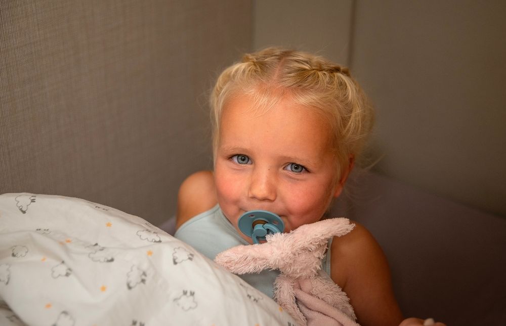 meisje (peuter) ligt in bed met speen en roze knuffeltje. dekbed heeft schaapjes patroon.