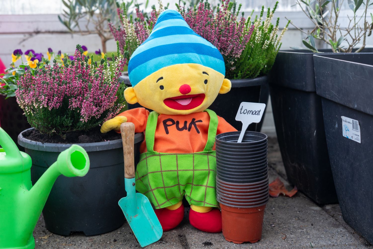 Knuffel staat met een schep naast planten. Knuffel is geel, heeft een blauw hoedje op, een oranje trui aan met 'Puk"erop en een groen tuinpakje aan.