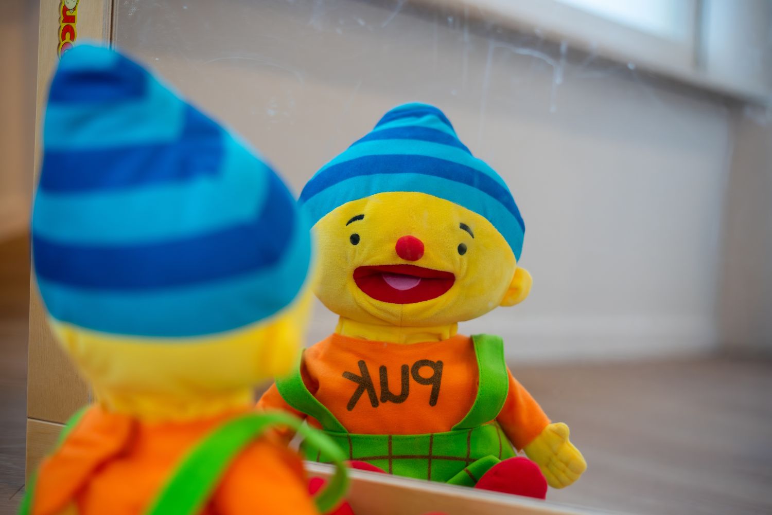 Knuffel kijkt in de spiegel. Knuffel is geel, heeft een blauw hoedje op, een oranje trui aan met 'Puk"erop en een groen tuinpakje aan. 