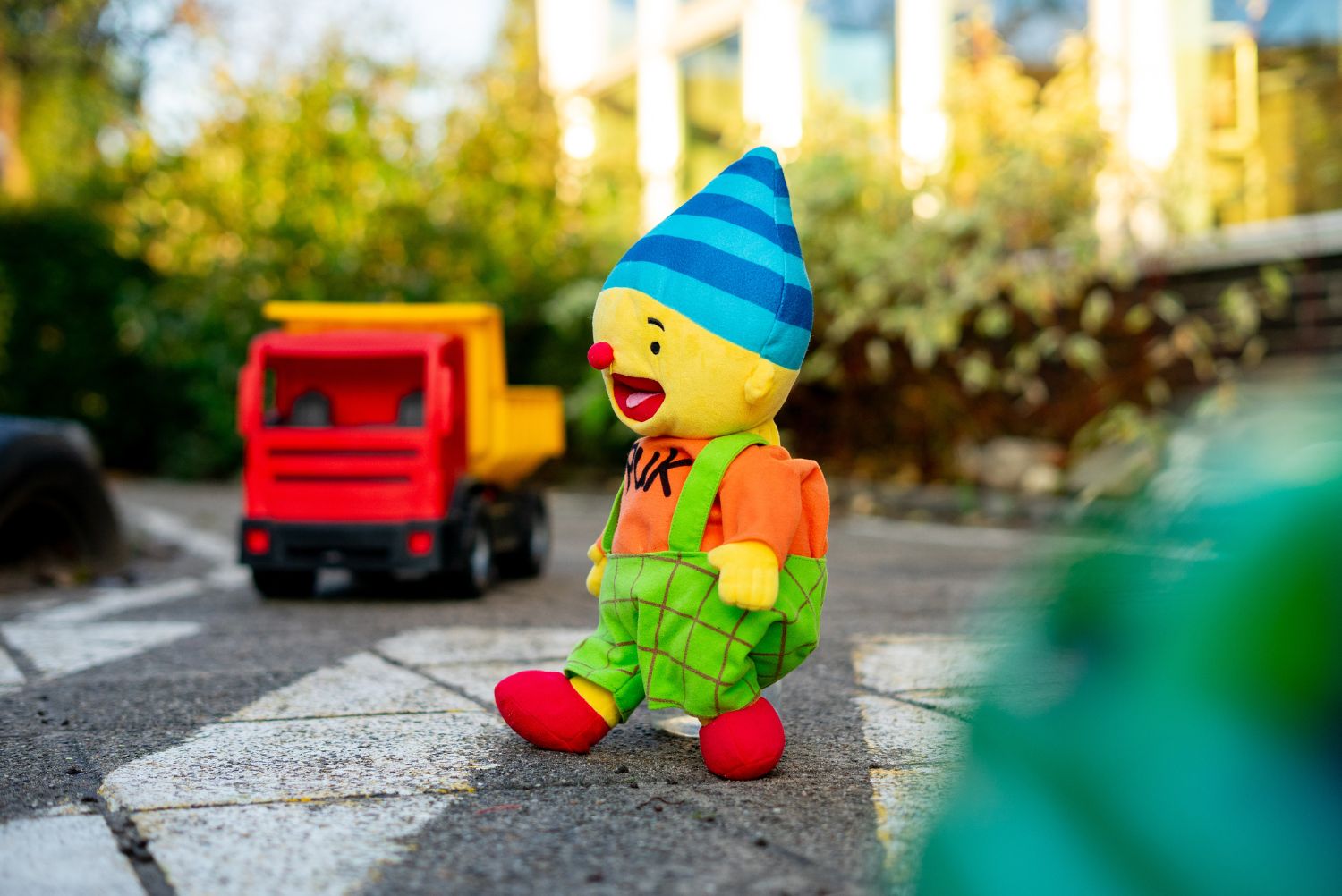Knuffel loopt op een zeebrapad. Daarachter staat een speelgoedvrachtwagen te wachten. Knuffel is geel, heeft een blauw hoedje op, een oranje trui aan met 'Puk"erop en een groen tuinpakje aan.