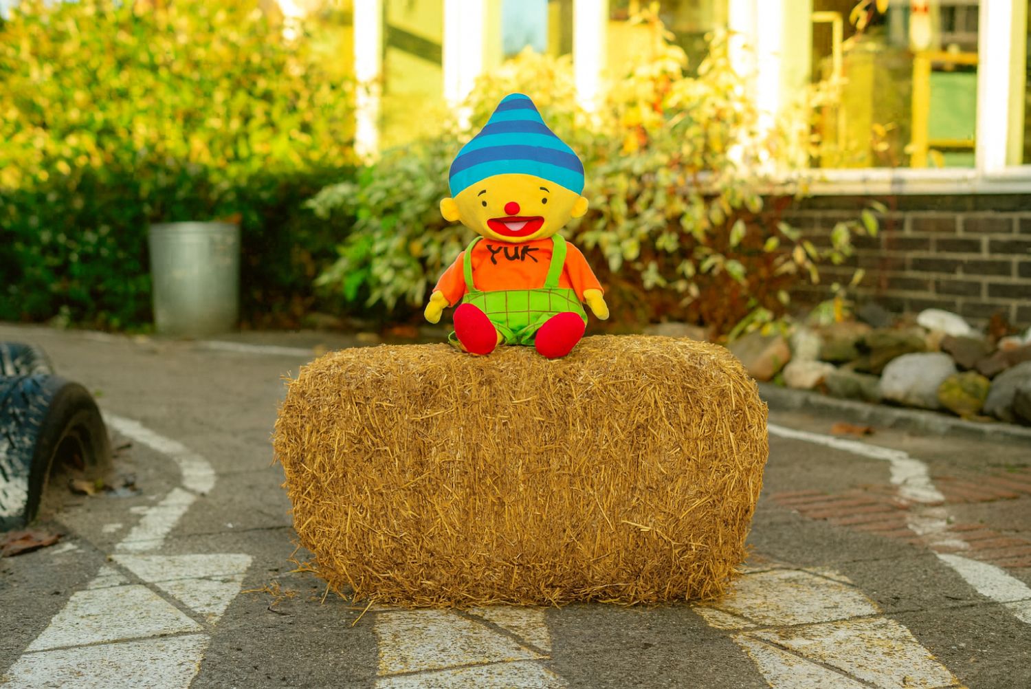 Knuffel zit op een hooibaal. Knuffel is geel, heeft een blauw hoedje op, een oranje trui aan met 'Puk"erop en een groen tuinpakje aan.